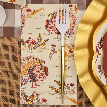 A gold and white fork rests on a Sophistiplate Amber Harvest Guest Towel, decorated with colorful turkeys and flowers, placed on a fall-themed table setting with gold-edged plates and a burlap and plaid tablecloth.